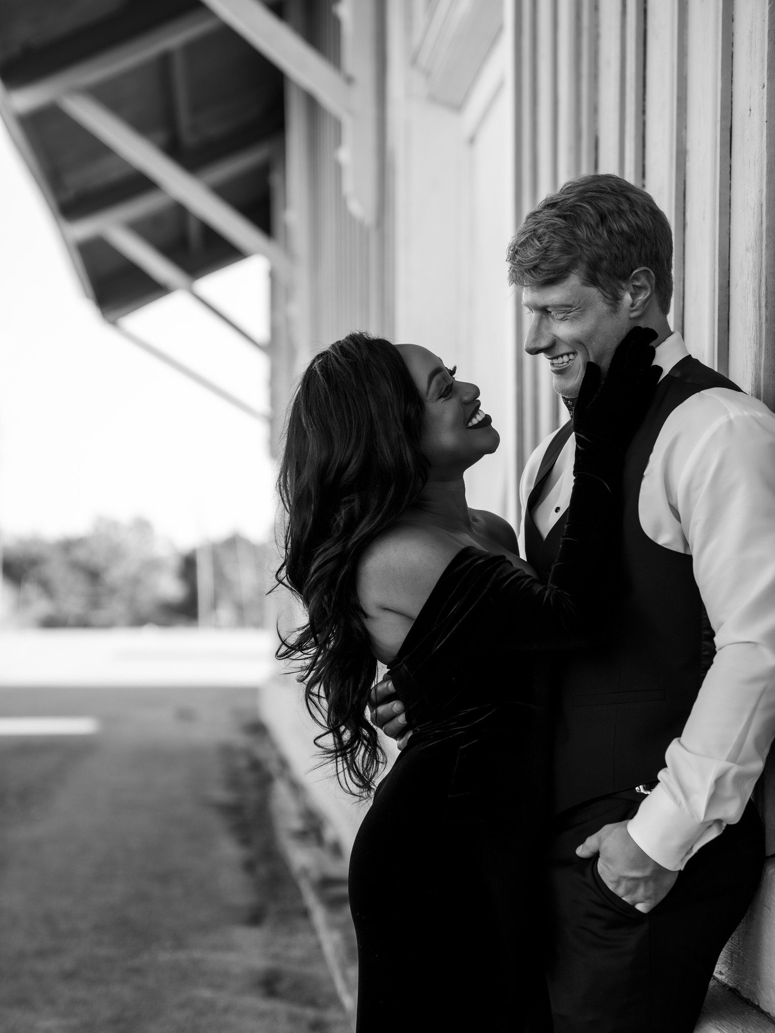 In black and white, Ashley laughs with her head tilted back while Jason smiles down at her beside the wooden building.