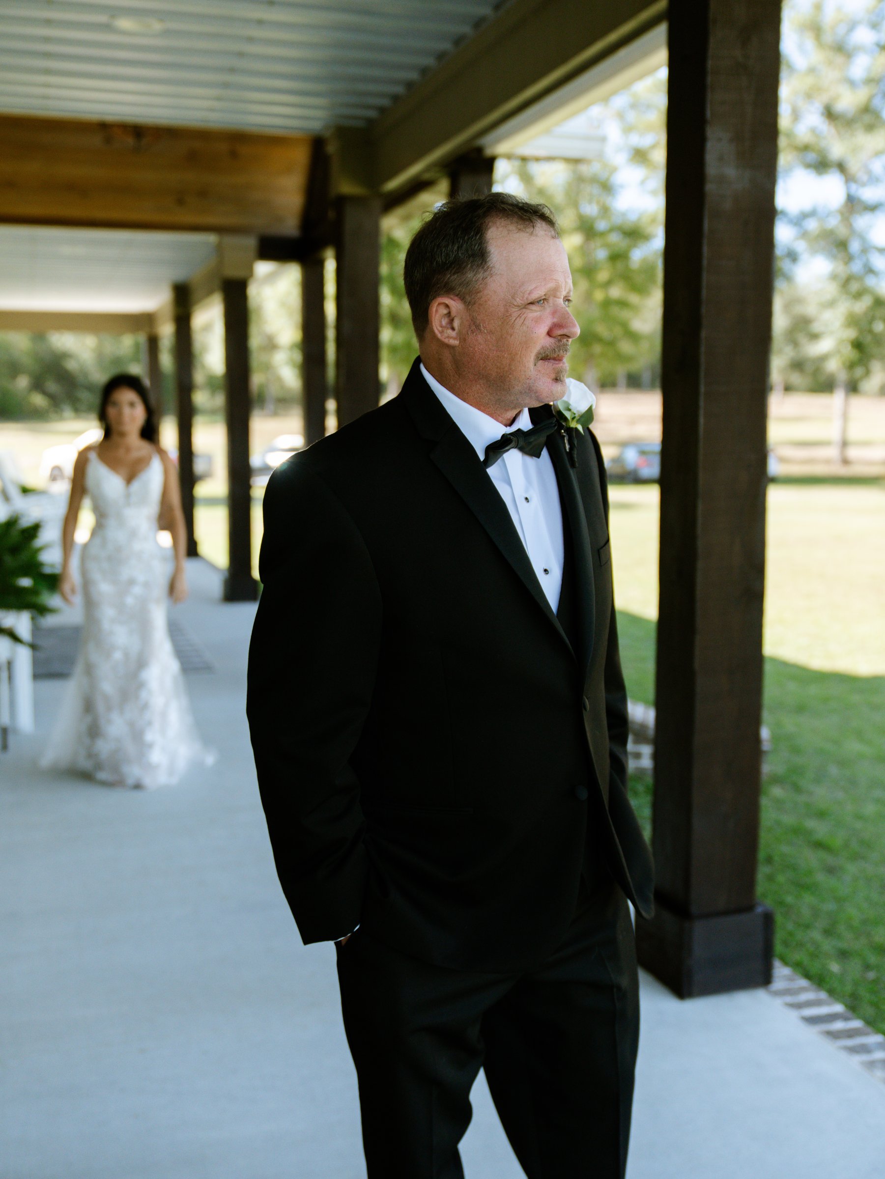 A man in a black tuxedo stands on a covered porch while Kenadee in her dress walks up behind him.