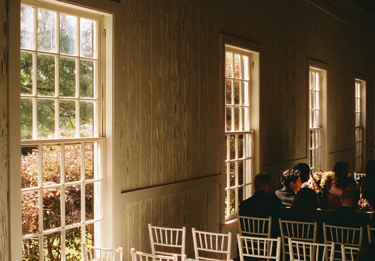 Sunlight through tall windows beside guests seated on white chairs at Emily and Timothy’s wedding, photographed on film.