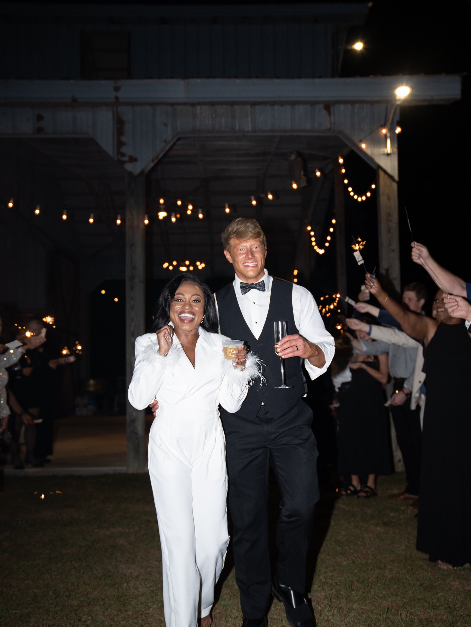 Ashley and Jason walk out arm in arm at night, smiling, with guests lining both sides and string lights overhead.