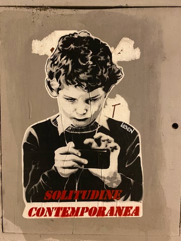 A large, high-contrast monochrome wheatpaste depicting a young boy absorbed in a smartphone. The artwork is accented with bold red typography at the bottom and is signed by the artist Laben.. Subject: A young boy with curly hair looking down at a mobile device.. Style: Stencil-style high-contrast monochrome with red stencil text.. Details: The boy is depicted in black and white with dramatic shadows, while the text 'SOLITUDINE CONTEMPORANEA' is printed in vibrant red at the base.. Shape: Die-cut silhouette. Setting: Urban environment. Surface: A flat urban surface, likely a utility cabinet or a painted door.. Colors: #000000 (Black), #FFFFFF (White), #B22222 (Firebrick Red), #8C8984 (Davy's Grey). Composition: Centered portrait orientation focusing on the subject's posture and the message at the bottom.. The piece provides a direct critique of modern digital addiction, particularly among the youth. The Italian phrase translates to 'Contemporary Solitude,' suggesting that while technology connects us, it simultaneously isolates individuals within a personal digital bubble, turning a social tool into a source of loneliness.
