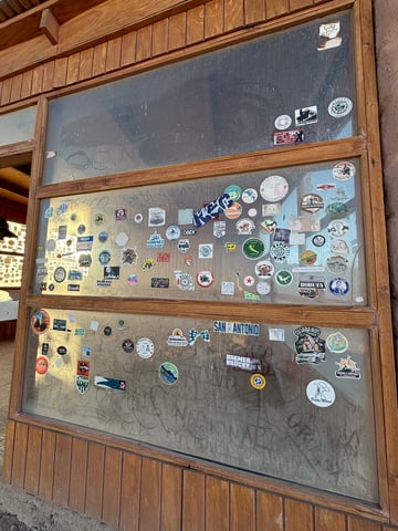 A large wooden-framed window divided into three horizontal glass panes is heavily adorned with a colorful mosaic of travel stickers. The decals represent a wide array of adventure-themed logos, motorcycle clubs, and 4x4 expedition markers, including references to 'ATACAMA 2019', 'MACHU PICCHU', and 'SAN ANTONIO'. The artistic styles are diverse, featuring circular badges, graphic illustrations, and bold typography. The stickers are a mix of vibrant colors—reds, blues, yellows, and greens—contrasting with the weathered, natural wood of the frame and the dusty, slightly scratched glass. This 'wall of fame' serves as a visual guestbook for travelers and adventurers, capturing a mood of wanderlust and shared exploration. The setting appears to be a rustic building, possibly a hostel or a shop in a remote travel hub, reflecting a community culture centered on road trips and South American discovery.
