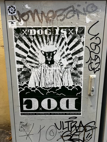 A large, high-contrast black and white wheatpaste poster dominates a weathered metal utility box. The artwork features a surreal, dog-headed figure in a messianic pose, surrounded by religious-style iconography and graphic text.. Subject: A humanoid figure with a dog's head wearing robes, positioned like a religious icon.. Style: Graphic, high-contrast illustration using halftone patterns and bold linework.. Details: The figure has outstretched hands atop a bed of clouds, with a sunburst 'halo' effect in the background. Text includes 'DOG IS' at the top and a large inverted 'DOG' at the bottom, with 'GU-TANG CLAN' repeated in the footer.. Shape: Rectangular poster. Setting: Urban environment. Surface: A metal electrical or utility cabinet located on a street.. Colors: #000000 (Black), #FFFFFF (White), #8C8C8C (Gray). Composition: Centered, eye-level frontal shot emphasizing the verticality of the poster.. This work subverts traditional religious imagery by replacing a holy figure with a canine, creating a juxtaposition between the sacred and the mundane. The inclusion of 'GU-TANG CLAN' serves as a playful homage to the Wu-Tang Clan, embedding the piece within hip-hop culture and urban mythology while questioning themes of devotion and public identity.