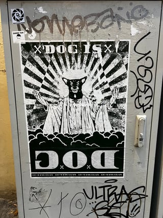 A large, high-contrast black and white wheatpaste poster dominates a weathered metal utility box. The artwork features a surreal, dog-headed figure in a messianic pose, surrounded by religious-style iconography and graphic text.. Subject: A humanoid figure with a dog's head wearing robes, positioned like a religious icon.. Style: Graphic, high-contrast illustration using halftone patterns and bold linework.. Details: The figure has outstretched hands atop a bed of clouds, with a sunburst 'halo' effect in the background. Text includes 'DOG IS' at the top and a large inverted 'DOG' at the bottom, with 'GU-TANG CLAN' repeated in the footer.. Shape: Rectangular poster. Setting: Urban environment. Surface: A metal electrical or utility cabinet located on a street.. Colors: #000000 (Black), #FFFFFF (White), #8C8C8C (Gray). Composition: Centered, eye-level frontal shot emphasizing the verticality of the poster.. This work subverts traditional religious imagery by replacing a holy figure with a canine, creating a juxtaposition between the sacred and the mundane. The inclusion of 'GU-TANG CLAN' serves as a playful homage to the Wu-Tang Clan, embedding the piece within hip-hop culture and urban mythology while questioning themes of devotion and public identity.