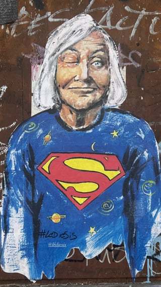 A large-scale wheatpaste artwork featuring an expressive portrait of an elderly woman winking, dressed in a cosmic-themed superhero garment. The piece is signed by the artist LeDiesis and is pasted onto a weathered wooden surface.. Subject: An elderly woman with white hair and a playful winking expression.. Style: Painterly illustration with bold brushwork and vibrant colors.. Details: She wears a blue sweater emblazoned with the iconic Superman 'S' logo, decorated with hand-drawn planets, stars, and swirls.. Shape: Die-cut portrait. Setting: Urban environment. Surface: A plywood construction hoarding or aged wooden wall with visible wood grain and old markings.. Colors: #2B67B2 (Cobalt Blue), #E21F26 (Heroic Red), #FEE101 (Bright Yellow), #F2F2F2 (Off-White), #583E31 (Earthy Brown). Composition: A centered, vertical portrait that focuses on the subject from the chest up, creating a direct connection with the viewer.. Part of the 'Superwomen' series by the artist LeDiesis, this work subverts traditional superhero imagery by casting an everyday elderly woman in the role of a hero. The wink suggests a playful 'secret' power, emphasizing that true heroism and strength can be found in wisdom, age, and maternal figures who have historically been overlooked in pop culture narratives.