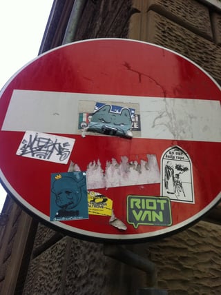 This image captures a 'No Entry' traffic sign heavily decorated with a variety of street art stickers, creating a layered collage of urban expression. The sign, a bright red circle with a white horizontal bar, serves as a canvas for several distinct pieces: a stylized blue-grey wolf or cat face, a black-and-white graffiti tag, a teal sticker featuring a cartoon character with a crown and wings captioned 'Dream Draw & Rocknroll!!!', and a lime-green 'RIOT VAN' sticker. Most notably, a provocative black-and-white sticker depicts a religious figure with the text '...give us our daily rape...', suggesting a strong political or social critique. The setting is a gritty urban environment, evidenced by the textured stone building in the background. The stickers are in various states of wear, with some peeling or torn, adding to the raw, underground aesthetic of the scene. This collection reflects themes of rebellion, subversion, and diverse artistic styles typical of sticker bombing and urban counter-culture.