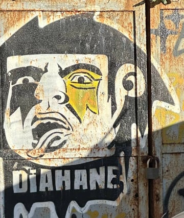 A large-scale high-contrast stencil of a stylized face with rock iconography applied to a heavily oxidized metal door. The artwork features a character with a star over one eye and a protruding tongue, accompanied by the text 'DIAHANE'.. Subject: A stylized human face reminiscent of glam rock icons, featuring a star over the eye.. Style: High-contrast graphic stencil art. Details: The piece includes the word 'DIAHANE' in a blocky font below the chin and uses yellow paint for the iconic star element.. Shape: Irregular die-cut style stencil. Setting: Urban industrial setting. Surface: A corrugated metal utility gate or door with a heavy padlock.. Colors: #A0522D (Rust Brown), #28282B (Matte Black), #F5F5F5 (Weathered White), #C5B358 (Muted Gold). Composition: Close-up, slightly angled shot that emphasizes the texture of the metal and the central graphic.. The piece merges the flashy aesthetics of 1970s glam rock with the gritty reality of industrial decay. By placing an icon of pop-culture stardom onto a rusted, locked gate, the artist creates a juxtaposition between fame and abandonment, or perhaps suggests that the spirit of rock persists even in the most neglected urban corners.