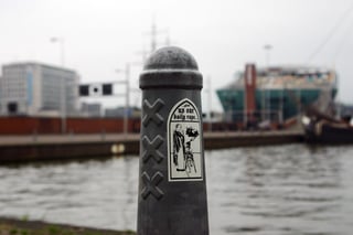 A provocative, black and white satirical sticker placed on a weathered grey Amsterdam bollard (Amsterdammertje) featuring the iconic triple St. Andrew's Crosses. The sticker is shaped like a gothic arched window, containing an illustration of a priest and a child on crutches. Above them, the text '...give us our daily rape...' parodies the Lord's Prayer, creating a biting critique of child abuse within the church. The setting is a waterfront in Amsterdam, with the distinctive green NEMO Science Museum and the masts of a ship blurred in the background. This piece of urban art utilizes dark humor and religious subversion as a form of political protest. The color palette is restricted to black ink on an off-white adhesive background.