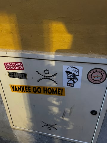 A collection of various stickers placed on a light grey metal utility cabinet set against a textured ochre wall. The scene is illuminated by strong, dappled sunlight casting organic shadows across the surface.