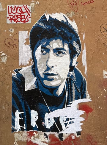 A weathered urban wall featuring a large, partially torn wheatpaste poster of a man's portrait in a stylized blue and white palette. Surrounding the poster are various hand-drawn tags, smaller stickers, and remnants of previous street art layers.