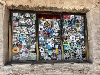 A dense and vibrant collection of hundreds of travel, motorcycle, and off-roading stickers layered over three glass window panes. This 'traveler's wall' features a chaotic yet organized collage of logos, graphic illustrations, and stenciled designs. Key visual elements include various club emblems like 'Ducati Official Club,' 'Poka Off Road,' and 'Rifredi Rude Boyz,' alongside geographical markers for places like Peru, Bolivia, Paraguay, and Zurich. The stickers vary in style from professional commercial prints to amateur artistic tags, using a full spectrum of colors—vibrant reds, deep blues, and bright yellows. The window is set within a thick, rustic wall made of textured, sandy-colored stone or mud brick, suggesting a remote waypoint, hostel, or roadside cafe. The overall mood is one of adventure and community, serving as a visual guestbook for global explorers and overlanders who have passed through this specific location.