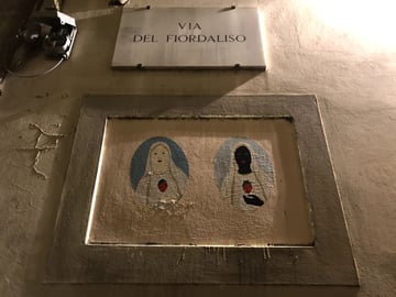 Two oval-shaped wheatpaste artworks are centered within a rectangular wall niche beneath a street sign in an urban setting at night. The pieces depict two religious-style figures in white robes, one with fair skin and the other with a dark, obscured face, both holding a red strawberry.. Subject: Two Madonna-like figures holding strawberries. Style: Modern illustrative iconography with flat colors. Details: Both figures wear white veils and robes against light blue backgrounds; the strawberry acts as a substitute for the traditional sacred heart.. Shape: Two separate elliptical (oval) wheatpastes. Setting: Urban night setting. Surface: A recessed rectangular niche in an exterior building wall. Colors: #F5F5DC (Beige), #FFFFFF (White), #000000 (Black), #ED1C24 (Strawberry Red), #A9C6D9 (Powder Blue). Composition: Centered, eye-level perspective focusing on the symmetry of the artwork within the architecture. The work subverts traditional religious iconography by introducing a duality of skin tone and replacing the 'Sacred Heart' with a strawberry. Located on 'Via del Fiordaliso' (Cornflower Street), the piece challenges historical European depictions of divinity, suggesting a more inclusive or culturally blended interpretation of the sacred in a modern urban context.