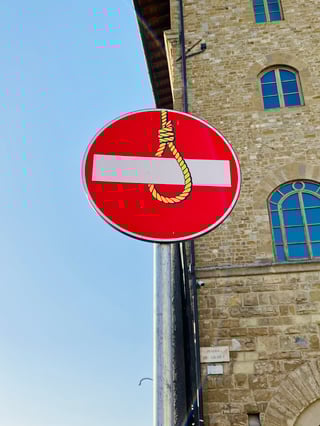 A circular 'No Entry' traffic sign has been creatively subverted with a yellow noose sticker. The artwork is positioned in front of a historic stone building under a clear blue sky.. Subject: A hangman's noose. Style: Graphic illustration. Details: Bright yellow rope with bold black outlines, cleverly interacting with the sign's horizontal white bar.. Shape: Die-cut. Setting: Historic urban square. Surface: Standard metal traffic sign on a utility pole. Colors: #E31E24 (Signal Red), #FFF200 (Safety Yellow), #F5F5F5 (Off-White), #87CEEB (Sky Blue), #A08C6B (Ancient Stone). Composition: Low-angle shot focusing on the sign, utilizing the building's corner to frame the right side.. This piece is a signature work of urban intervention, transforming a symbol of bureaucratic command into a metaphor for the lethal or stifling nature of absolute restriction. By placing the noose around the 'No Entry' bar, the artist suggests that blind obedience to boundaries can lead to a figurative social death, inviting viewers to question the rules that govern their movement and behavior.