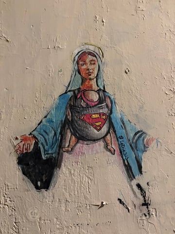 This street art features a contemporary rendition of a Madonna-like figure winking playfully while carrying a baby in a carrier. The infant's carrier is decorated with the iconic Superman 'S' shield, blending religious imagery with modern pop culture.. Subject: A modern Madonna figure winking and carrying a baby. Style: Illustrative and painterly with a stencil-like influence. Details: The figure wears a traditional blue cloak and has a halo, but the addition of a baby carrier with a Superman logo and the wink creates a humorous, empowering contrast.. Shape: Irregular die-cut. Setting: Urban environment. Surface: A roughly plastered urban wall. Colors: #ADD8E6 (Sky Blue), #E23636 (Classic Red), #FFD700 (Golden Yellow), #F5F5DC (Beige), #000000 (Charcoal Black). Composition: Centered portrait view emphasizing the figure's expression and the symbolic carrier.. Created by the artist Lediesis, this piece is part of a series that 'unmasks' female icons to reveal their inner strength. By merging the sacred Virgin Mary with the secular Superman symbol, the artist celebrates the divinity and heroism inherent in motherhood and the everyday woman, using the 'wink' to subvert traditional, passive religious tropes.