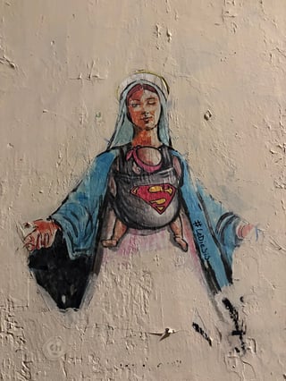 This street art features a contemporary rendition of a Madonna-like figure winking playfully while carrying a baby in a carrier. The infant's carrier is decorated with the iconic Superman 'S' shield, blending religious imagery with modern pop culture.. Subject: A modern Madonna figure winking and carrying a baby. Style: Illustrative and painterly with a stencil-like influence. Details: The figure wears a traditional blue cloak and has a halo, but the addition of a baby carrier with a Superman logo and the wink creates a humorous, empowering contrast.. Shape: Irregular die-cut. Setting: Urban environment. Surface: A roughly plastered urban wall. Colors: #ADD8E6 (Sky Blue), #E23636 (Classic Red), #FFD700 (Golden Yellow), #F5F5DC (Beige), #000000 (Charcoal Black). Composition: Centered portrait view emphasizing the figure's expression and the symbolic carrier.. Created by the artist Lediesis, this piece is part of a series that 'unmasks' female icons to reveal their inner strength. By merging the sacred Virgin Mary with the secular Superman symbol, the artist celebrates the divinity and heroism inherent in motherhood and the everyday woman, using the 'wink' to subvert traditional, passive religious tropes.