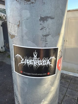 A horizontal rectangular sticker for the musical project 'Livergga', prominently displayed on a weathered, grey metallic utility pole in an urban outdoor setting. The sticker features a classic black metal aesthetic, utilizing a stark black background with a white, highly stylized, and spiky logo that appears to drip like icicles or blood. An inverted cross is integrated into the central part of the typography. Below the main band name, the web address 'livergga.bandcamp.com' is written in a clean white font, flanked by two small, intricate red circular symbols that resemble chaos stars. The pole itself shows signs of age with peeling paint and scratches. The background depicts a sidewalk with textured tiles and a glimpse of a gate, suggesting a residential or city street. The overall mood is dark, aggressive, and representative of underground extreme metal subculture.