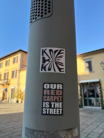 Two distinct stickers are positioned vertically on a smooth, gray utility pole in a Mediterranean-style urban square. The composition captures the contrast between modern street culture and traditional architecture under a clear blue sky.. Subject: Bold typographic message and a stylized logo tag. Style: Graphic design and block typography. Details: The lower sticker features the slogan 'OUR RED CARPET IS THE STREET' in bold white sans-serif font on a dark background. Above it, a square sticker features a distorted radial 'KOTE' design with pink and green chromatic aberration effects.. Shape: rectangular. Setting: Urban European square. Surface: A modern cylindrical metal utility or light pole. Colors: #8C8C8C (Cool Gray), #FFFFFF (Pure White), #000000 (Jet Black), #E8A28C (Building Ochre), #87CEEB (Sky Blue). Composition: A low-angle vertical shot that emphasizes the height of the pole against the sky.. The stickers represent a claim to public space, with the lower slogan explicitly elevating the grit of the street to the status of a 'red carpet'. This reflects a common theme in skate and street culture where the urban environment is viewed as a stage for personal expression and performance.
