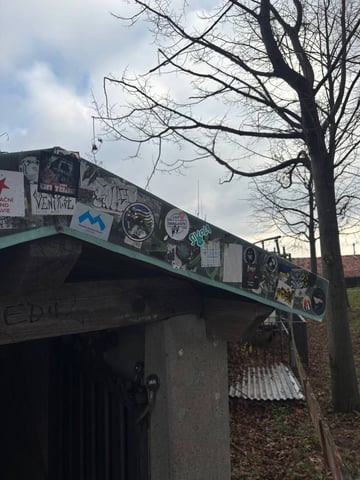 A diverse array of street stickers layered along the green metal edge of a wooden shelter's roof. This collection features an eclectic mix of graphic designs, including logos, subcultural emblems, and typography. Notable stickers include a 'MUNICHANDUTEAM' logo with a blue 'M', 'MotoLocura', and 'SIVOLA' in stylized green font, alongside several 'ULTRAS' football-related motifs with eagles and yellow-black color schemes. The stickers are placed on a weathered outdoor structure made of rough wood and concrete, situated in an environment with bare trees and a cloudy sky. The collection represents a visual record of travelers and subcultural groups marking their presence. Visible text such as 'On Tour', a red star sticker with partial Czech text, and hand-carved 'EDI' in the wood below, add to the layered narrative of the site. The mood is one of urban subculture meeting nature, showcasing the common practice of 'sticker slapping' at public rest stops.
