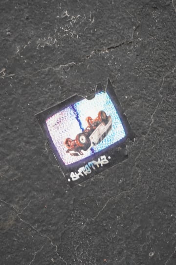 A weathered, rectangular sticker featuring a pixelated image of an overturned car, placed on a dark, cracked asphalt-like surface. The artwork has a nostalgic, low-fidelity aesthetic reminiscent of vintage television static or a surveillance freeze-frame.. Subject: An overturned vehicle. Style: Lo-fi glitch art / Pixelated. Details: The car is depicted in high-contrast white and rust tones against a blue-tinted static background. A stylized white tag reading '.SHTOTHS.' sits at the bottom.. Shape: Rectangular with a small tear at the top edge. Setting: Urban exterior. Surface: Asphalt pavement or a heavily textured concrete wall. Colors: #2E2E2E (Charcoal), #FFFFFF (Signal White), #7FB3D5 (Glitch Blue), #C0392B (Rust Red). Composition: Top-down perspective focusing on the central placement of the sticker. The piece evokes a sense of 'urban voyeurism' and digital nostalgia. By rendering a car crash through the lens of low-resolution static, it creates an emotional distance, transforming a moment of chaos into a fixed aesthetic signal. Its placement on a gritty, road-like surface grounds the digital glitch in a physical reality, suggesting a literal and metaphorical 'breakdown' in the urban fabric.