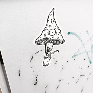 This black and white die-cut sticker features a grotesque, psychedelic mushroom monster. The tall, tapering cap is covered in numerous unblinking eyeballs of various sizes, with a large, prominent eye at its center. Its stalk is transformed into a screaming face with a wide mouth full of needle-like teeth and a long, slender tongue lashing out. The illustration is rendered in a detailed ink-sketch style with fine hatching and stippling, typical of underground comic or counter-culture art. The sticker is adhered to a white, textured metal or plastic surface, possibly a utility box, which shows minor signs of wear with black scuffs and a faint, pale green graffiti tag to the right. The piece carries a surreal, trippy, and slightly dark mood, referencing psychedelic experiences and monstrous transformations.