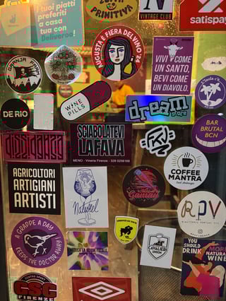 A dense and vibrant collage of various stickers covering a transparent glass window. The collection features a mix of local business logos, social messages, and artistic illustrations, creating a textured urban tapestry.. Subject: Artistic representation of a wine glass. Style: Line art illustration. Details: A tall, elegant wine glass containing a stylized female figure with flowing hair, accompanied by the word 'Naturel' in a flowing cursive script.. Shape: Rectangular. Setting: Indoor urban establishment. Surface: Window or door of a bar or restaurant. Colors: #800020 (Burgundy), #FFFFFF (White), #000000 (Black), #4B0082 (Indigo), #FFD700 (Gold). Composition: A flat-lay perspective focusing on the density and overlapping layers of the sticker collection.. The collage acts as a communal canvas, documenting the cultural and commercial identity of the local neighborhood. It blends themes of gastronomy, social activism (e.g., 'Free Palestine'), and personal artistic expression, turning a functional glass surface into a vibrant piece of public history.