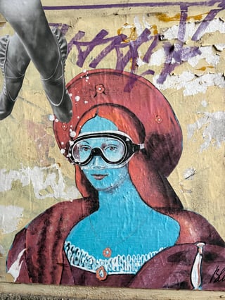 A large-scale surrealist wheatpaste mural featuring a blue-skinned Renaissance-style woman wearing diving goggles. The artwork is layered over a weathered urban wall with visible remnants of previous graffiti and peeling paint.. Subject: A woman in classical portraiture style with blue skin and diving goggles. Style: Surrealist collage blending Renaissance aesthetic with modern objects. Details: The figure has vibrant turquoise skin, a deep red circular headpiece, and is depicted with bubbles near her face. Grayscale photographic ballet feet are pasted in the upper left corner.. Shape: Large irregular wheatpaste cutout. Setting: Urban environment. Surface: An urban exterior wall. Colors: #40B5D1 (Turquoise Blue), #A33E43 (Burgundy Red), #E5D7A1 (Pale Yellow), #7E549F (Grape Purple), #FFFFFF (White). Composition: A centered portrait composition with a vertical orientation that captures the interaction between the art and the decaying wall.. This piece utilizes juxtaposition to create a dreamlike narrative, blending the high-culture aesthetics of the Renaissance and ballet with modern diving gear. The blue skin and goggles suggest a theme of exploration or preservation within a submerged, alternative reality, while its placement on a decaying urban wall highlights the contrast between timeless art and the ephemeral nature of street culture.