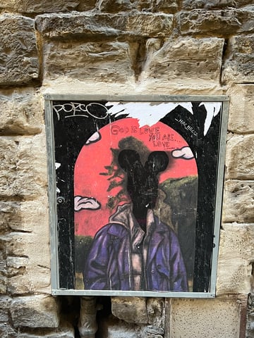 A large, illustrative street art poster is displayed within a metal frame recessed into a weathered stone wall. The artwork features a silhouetted figure with iconic rounded ears against a vibrant pink sky, accompanied by hand-written spiritual text.. Subject: A silhouetted figure in a purple jacket and hoodie with mouse-like ears.. Style: Painterly illustration with street art tagging.. Details: The text 'GOD IS LOVE YOU ARE... LOVE' is written in a thin, marker-like font. There are white clouds and a landscape in the background of the inner arch.. Shape: rectangular. Setting: Urban exterior. Surface: A metal utility box or display frame embedded in a masonry wall.. Colors: #4B3B7A (Royal Purple), #EB6B7F (Salmon Pink), #1A1A1A (Carbon Black), #A89F91 (Stone Tan). Composition: Straight-on, centered shot that emphasizes the contrast between the colorful art and the monochromatic stone.. The work juxtaposes spiritual affirmations with pop-culture iconography and the grittiness of urban decay. By blacking out the face, the artist creates a universal figure that represents humanity's search for identity and divinity within a commercialized world.