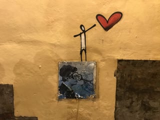 A mixed-media urban artwork featuring a central sticker of a kissing couple integrated with hand-drawn elements on a warm yellow wall. Above the sticker, a minimalist stick figure reaches toward a vibrant red heart, creating a narrative of affection.. Subject: Two people wearing vintage-style goggles sharing a kiss. Style: illustrated sketch. Details: Includes handwritten text 'Steve <3 Mo 2017' and has a blue-toned monochromatic color scheme.. Shape: square. Setting: urban environment. Surface: exterior urban wall with chipped plaster. Colors: #E4C57F (Ochre Yellow), #3E5C8D (Steel Blue), #BD2D2D (Deep Red), #000000 (Ink Black). Composition: Centered focal point on the sticker with vertical movement leading to the hand-drawn heart.. The piece explores the intersection of personal memory and public space. By combining a realistic, blue-toned depiction of a specific couple with a minimalist stick figure, the artist transforms a private romantic moment into a universal symbol of love's endurance within the decaying urban landscape.