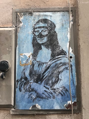 A large, weathered wheatpaste poster on a metal utility door depicting a stylized Mona Lisa wearing a diving mask. The artwork features a monochromatic blue background and show signs of environmental aging like peeling and cobwebs.. Subject: The Mona Lisa wearing a snorkel mask. Style: stencil-influenced illustrative sketch. Details: The iconic figure is rendered in black ink against a textured blue backdrop, with a detailed black diving mask covering the eyes and nose.. Shape: rectangular. Setting: urban exterior. Surface: a recessed metal utility box door set into a stone or stucco building wall. Colors: #89B6D2 (Sky Blue), #1A1A1B (Charcoal Black), #C5C0B8 (Concrete Grey), #D4C2A1 (Aged Paper). Composition: straight-on eye-level shot. This piece subverts a high-culture icon by placing it in a mundane urban context and adding an absurd modern accessory. The diving mask may suggest themes of environmental preservation or the idea of classical art 'drowning' or needing to adapt to a changing modern landscape.