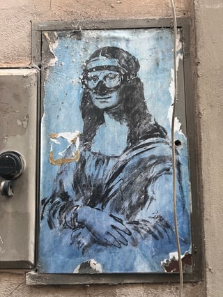 A large, weathered wheatpaste poster on a metal utility door depicting a stylized Mona Lisa wearing a diving mask. The artwork features a monochromatic blue background and show signs of environmental aging like peeling and cobwebs.. Subject: The Mona Lisa wearing a snorkel mask. Style: stencil-influenced illustrative sketch. Details: The iconic figure is rendered in black ink against a textured blue backdrop, with a detailed black diving mask covering the eyes and nose.. Shape: rectangular. Setting: urban exterior. Surface: a recessed metal utility box door set into a stone or stucco building wall. Colors: #89B6D2 (Sky Blue), #1A1A1B (Charcoal Black), #C5C0B8 (Concrete Grey), #D4C2A1 (Aged Paper). Composition: straight-on eye-level shot. This piece subverts a high-culture icon by placing it in a mundane urban context and adding an absurd modern accessory. The diving mask may suggest themes of environmental preservation or the idea of classical art 'drowning' or needing to adapt to a changing modern landscape.