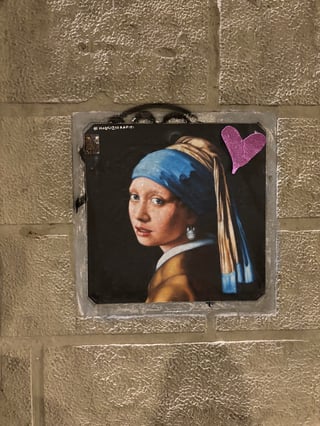 A playful street art parody of Johannes Vermeer's 'Girl with a Pearl Earring', where the subject's iconic pearl is replaced by a tiny, white Totoro character from Studio Ghibli. The artwork is a square paste-up or tile mounted onto a textured, light-colored stone block wall. The illustration maintains a classical painterly aesthetic with a vibrant blue headscarf, a golden-ochre jacket, and a deep black background. A small, sparkling pink glitter heart sticker is adhered to the top right corner of the piece. The hashtag '#MAURIZIORAPITI' is visible in small white lettering at the top left edge. This piece represents a whimsical mashup of high art and modern pop culture, adding a touch of surrealism and humor to an urban environment.