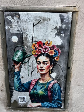A vibrant wheatpaste of a woman inspired by Frida Kahlo, adorned with a colorful floral crown and holding a green watering can. The artwork is applied to a weathered metal utility panel set within a textured urban wall.. Subject: Frida Kahlo-inspired figure with a floral headdress. Style: painterly illustration. Details: The figure holds a green watering can labeled 'MISS QUARK' and wears traditional jewelry; a QR code is visible at the bottom left.. Shape: die-cut wheatpaste. Setting: urban streetscape. Surface: utility access door on an exterior wall. Colors: #C0392B (Crimson Red), #2C3E50 (Midnight Blue), #27AE60 (Emerald Green), #F39C12 (Orange), #BDC3C7 (Silver Gray). Composition: centered, eye-level portrait framing. This piece recontextualizes the image of Frida Kahlo as a symbol of growth and resilience. By depicting her with a watering can, the artist suggests that creativity and cultural heritage require constant nurturing to thrive within the harsh, gray environment of the city.