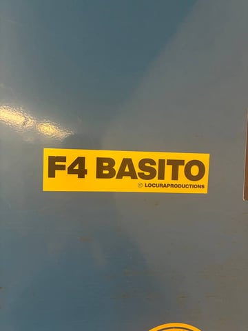 A minimalist rectangular sticker featuring high-contrast graphic design. The sticker has a bright yellow background with bold, black sans-serif typography that reads 'F4 BASITO'. Below the main text, in much smaller lettering, is the handle '@ LOCURAPRODUCTIONS' accompanied by an Instagram icon. The sticker is applied to a smooth, matte blue surface, likely a piece of street furniture like a metal utility box or a locker. The overall style is clean and promotional, typical of urban tagging and branding. The stark color palette of yellow, black, and the blue background creates a strong visual impact, making it easily identifiable in an urban setting. This piece reflects contemporary street-level marketing and collective identity.