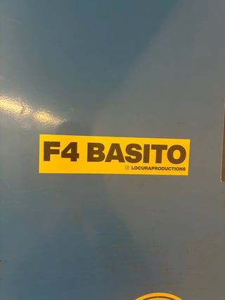 A minimalist rectangular sticker featuring high-contrast graphic design. The sticker has a bright yellow background with bold, black sans-serif typography that reads 'F4 BASITO'. Below the main text, in much smaller lettering, is the handle '@ LOCURAPRODUCTIONS' accompanied by an Instagram icon. The sticker is applied to a smooth, matte blue surface, likely a piece of street furniture like a metal utility box or a locker. The overall style is clean and promotional, typical of urban tagging and branding. The stark color palette of yellow, black, and the blue background creates a strong visual impact, making it easily identifiable in an urban setting. This piece reflects contemporary street-level marketing and collective identity.