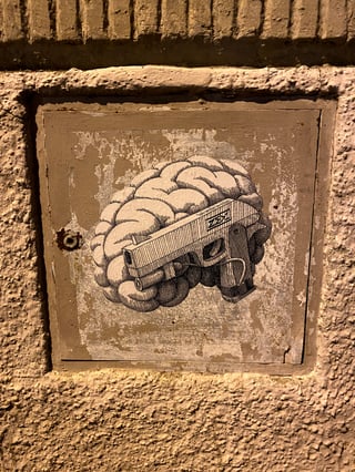 A black and white line-art sticker depicting a human brain fused with a handgun, placed on a weathered metal utility hatch. The hatch is embedded in a rough, beige-colored stucco or concrete wall.. Subject: A human brain integrated with a semi-automatic handgun.. Style: Detailed black and white illustration with cross-hatching and fine-line shading.. Details: The gun's slide and grip are intricately merged into the folds of the brain, featuring a small 'ZEMS' tag on the side.. Shape: Die-cut. Setting: Urban exterior. Surface: A metal utility hatch or electrical box door set into a building exterior.. Colors: #968576 (Muted Brown), #D2B48C (Tan), #000000 (Black), #FFFFFF (White), #705E52 (Umber). Composition: Centered, close-up shot focusing on the sticker within the frame of the metal box.. The piece explores the intersection of thought and violence, suggesting that ideas can be as lethal as physical weapons. By merging an organ associated with consciousness with a tool of destruction, the artist comments on the weaponization of ideology within the urban environment.