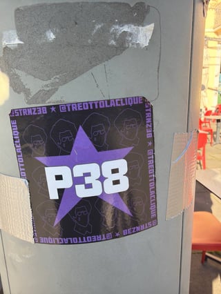 A rectangular graphic sticker is prominently displayed on a weathered metal utility pole. The sticker features bold text and a star motif, reinforced by silver duct tape on its sides.. Subject: The text 'P38' superimposed on a purple star with repeating line-art portraits in the background.. Style: Graphic vector illustration with a focus on bold typography.. Details: The sticker includes social media handles around the border and features stylized line-drawn faces of a man with an afro and glasses.. Shape: rectangular. Setting: Urban outdoor environment. Surface: A cylindrical metallic utility pole or street light post.. Colors: #8A2BE2 (Violet Star), #FFFFFF (White Typography), #000000 (Black Background), #808080 (Grey Metal Pole). Composition: Close-up, centered on the sticker with a shallow depth of field that blurs the street scene behind.. The sticker appears to be promotional material for a musical collective or underground culture movement. By utilizing a five-pointed star and bold alphanumeric codes, it adopts a visual language of political rebellion or 'vanguard' status, re-contextualizing these symbols for modern urban self-promotion.
