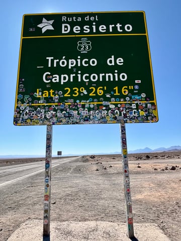 A large green road sign marking the Tropic of Capricorn is heavily adorned with hundreds of stickers from travelers. The sign stands prominently against a vast, arid desert landscape under a brilliant blue sky.. Subject: Traveler tags, regional flags, and club logos. Style: graphic and illustrative. Details: A dense, overlapping mosaic of 'slaps' that completely cover the lower portion of the sign face and its support columns.. Shape: mixed (circular, rectangular, and die-cut). Setting: remote desert. Surface: large highway landmark sign. Colors: #004D2C (Forest Green), #F9D308 (Bright Yellow), #8DB8E0 (Sky Blue), #C4B7A5 (Desert Sand). Composition: Centered, wide-angle shot highlighting the scale of the sign against the horizon. The stickers transform a functional geographical marker into a collaborative monument of global pilgrimage. Each sticker serves as a digital-age 'Kilroy was here', representing a collective memory of travelers who have crossed this specific point on the Earth's surface.