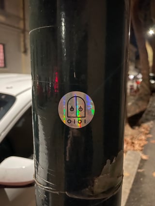 A minimalist circular holographic sticker is centered on a dark, glossy utility pole. The sticker features black graphic lines depicting an arched doorway with power-button symbols, set against a shimmering iridescent background.