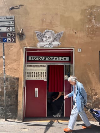 A large black-and-white cherub wheatpaste is positioned directly above a vintage red 'Fotoautomatica' booth on a textured ochre wall. In the foreground, an elderly woman with a cane and a shopping trolley walks past, creating a scene of everyday urban life.. Subject: A classical Renaissance-style cherub with wings, resting its head on its hands.. Style: Illustrated greyscale with fine-line shading.. Details: The figure features a speech bubble at the bottom containing the colloquial Tuscan phrase 'BOIA DHE'.. Shape: Die-cut wheatpaste. Setting: Urban streetscape. Surface: The exterior wall of a building in an Italian street, likely Florence given the signage.. Colors: #D2A679 (Ochre), #A31F24 (Crimson), #FFFFFF (White), #2E2E2E (Charcoal). Composition: A vertical shot utilizing the rule of thirds, with the photo booth and cherub as the central anchor.. The juxtaposition of the angelic figure over a machine designed to capture human identity (the photo booth) suggests a dialogue between the eternal and the ephemeral. By using a Renaissance motif in a modern street context, the artist bridges the gap between Italy's high-art history and contemporary urban vernacular.