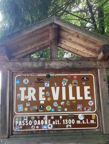 A rustic wooden information sign for Passo Daone at an altitude of 1300 meters, heavily adorned with a diverse collection of travel and club stickers. The sign is framed by heavy timber and set against a backdrop of lush green forest foliage.. Subject: The 'Mistoerer' graphic logo. Style: minimalist graphic. Details: Features bold black typography alongside a silhouette of a person engaged in outdoor sports, characteristic of travel tag stickers.. Shape: rectangular. Setting: alpine forest. Surface: A metal information board mounted inside a protective wooden roofed structure.. Colors: #72412C (Sign Brown), #7D5A44 (Wood Bark), #FFFFFF (Stark White), #2F411C (Deep Forest Green). Composition: A centered, eye-level shot that captures the sign as a repository of collective memory.. The sign serves as a 'living' archive of human passage through the landscape. By placing stickers on a functional piece of infrastructure, travelers transform a bureaucratic marker into a communal canvas that records their presence and shared passions for the outdoors.