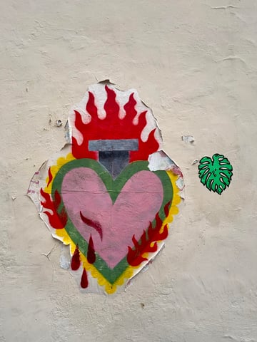 A large, colorful wheatpaste of a stylized sacred heart dominates the center of a textured wall, showing visible signs of weathering and peeling. Accompanying it is a smaller, vibrant green monstera leaf sticker, creating a juxtaposition between religious iconography and modern botanical trends.. Subject: A stylized Sacred Heart with flames and a dagger hilt. Style: Graphic illustration inspired by religious iconography. Details: The heart features a pink center with a green border and yellow scalloped edges, topped with vibrant red flames and three falling blood droplets at the base.. Shape: Large irregular die-cut wheatpaste. Setting: Urban street environment. Surface: External urban plastered wall. Colors: #FF2400 (Scarlet Red), #FFB6C1 (Light Pink), #F9D71C (Bright Yellow), #2E8B57 (Sea Green), #E5E4E2 (Platinum Beige). Composition: Centered main subject with a smaller secondary element positioned to the right to create visual balance. This work repurposes the Sacred Heart—a traditional symbol of divine devotion and suffering—into a modern street art context. The peeling paper reflects the ephemeral nature of street art and the inevitable decay of passion over time, while the adjacent Monstera sticker adds a contemporary, secular touch of 'urban jungle' aesthetic to the composition.