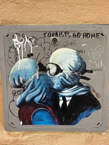 A street art wheatpaste depicting two hooded figures in an intimate kiss while wearing diving goggles, placed on a weathered utility panel. The piece is a modern urban reinterpretation of René Magritte's 'The Lovers II'.. Subject: Two figures embracing and kissing with their heads covered in blue fabric and wearing scuba diving masks.. Style: Painterly illustrative street art. Details: Includes the artist's signature 'Blub' at the bottom and features detailed fabric folds and light reflections on the goggles.. Shape: die-cut. Setting: urban building exterior. Surface: A metal or plastic utility access box embedded in an exterior wall.. Colors: #315A8E (Deep Blue), #8E8E8E (Grey), #CBA266 (Ochre), #1A1A1A (Black), #7E2328 (Burgundy). Composition: Centered, eye-level medium shot focusing on the interaction between the street art and the surrounding graffiti.. Part of the 'L'arte sa nuotare' series, this piece suggests that art and human emotion can survive and 'swim' even during times of crisis or cultural saturation. The juxtaposition with the 'Tourists Go Home' tag reflects the tension between global art icons and the local realities of over-tourism in historic cities.