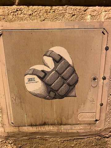 A large black and white illustrated sticker of a heart encased in tactical gear, pasted onto a ribbed metal utility box. The artwork combines soft anatomical shapes with the rigid structure of a bulletproof vest, accompanied by Italian text.. Subject: A heart wearing a tactical plate carrier/bulletproof vest. Style: Hatched ink illustration. Details: High-contrast black ink with intricate cross-hatching for depth, featuring the tag 'ZEM' and the phrase 'Quel cuore da proteggere' (That heart to protect).. Shape: Heart-shaped die-cut. Setting: Urban evening. Surface: A ribbed Enel utility box set into a rough-textured urban wall.. Colors: #000000 (Onyx), #FFFFFF (Cloud), #C6B6A5 (Almond), #8B7E72 (Taupe). Composition: Centered, straight-on medium shot highlighting the juxtaposition of the art and the industrial texture.. This piece explores themes of emotional vulnerability and the defensive mechanisms developed in response to urban or personal conflict. By cladding the universal symbol of love in combat gear, the artist 'ZEM' comments on the necessity of protecting one's inner sensitivity in a world that often feels hostile or demanding.