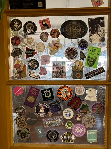 A dense and diverse collection of urban stickers layered onto two glass window panes separated by a yellow wooden frame. The collection reflects a wide array of themes including psychedelic art with glowing mushrooms, cosmic eyes, and intricate sacred geometry mandalas; outdoor culture featuring cacti, mountains, and trees; and local branding for music systems and artisanal products. Notable subjects include a floating Tanooki Mario illustration, a neon green 'Poison Griptape' bottle, and a purple skull wearing a bucket hat. The artistic styles range from detailed digital illustrations and bold typography to photographic elements and stencil-like graphics. Visible text includes phrases such as 'Natural Radio', 'Mad Charlie's', 'SOMA Mushroom Elixir', 'Chakano Sound System', and 'Ecoterapia'. The overall mood is eclectic and vibrant, suggesting a bohemian setting like a cafe, skate shop, or creative studio where community members have layered their personal and brand identities over time.