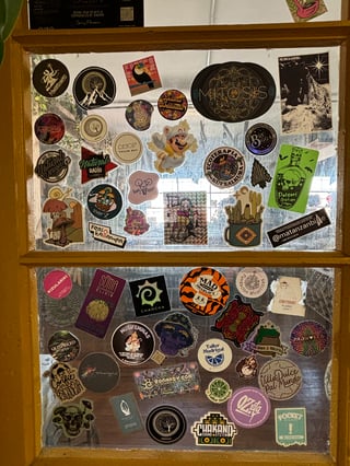 A dense and diverse collection of urban stickers layered onto two glass window panes separated by a yellow wooden frame. The collection reflects a wide array of themes including psychedelic art with glowing mushrooms, cosmic eyes, and intricate sacred geometry mandalas; outdoor culture featuring cacti, mountains, and trees; and local branding for music systems and artisanal products. Notable subjects include a floating Tanooki Mario illustration, a neon green 'Poison Griptape' bottle, and a purple skull wearing a bucket hat. The artistic styles range from detailed digital illustrations and bold typography to photographic elements and stencil-like graphics. Visible text includes phrases such as 'Natural Radio', 'Mad Charlie's', 'SOMA Mushroom Elixir', 'Chakano Sound System', and 'Ecoterapia'. The overall mood is eclectic and vibrant, suggesting a bohemian setting like a cafe, skate shop, or creative studio where community members have layered their personal and brand identities over time.