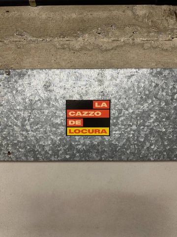 A graphic, typographic street sticker featuring the phrase "LA CAZZO DE LOCURA" in a bold, sans-serif white font. The text is arranged in stacked, color-coded blocks of red and yellow against a black rectangular background. The phrase is a multilingual mashup of Italian and Spanish slang, translating roughly to "the fucking madness". The sticker is placed in the center of a galvanized metal plate that has a textured, crystalline pattern. This metal plate is affixed to a wall, sandwiched between a rough concrete upper section and a smooth, light-grey lower section. The overall aesthetic is industrial and minimalist, reflecting an urban environment. The piece captures a mood of frustration, energy, or a cynical commentary on modern life through bold linguistic play.