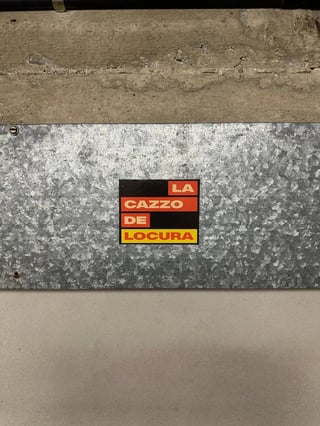 A graphic, typographic street sticker featuring the phrase "LA CAZZO DE LOCURA" in a bold, sans-serif white font. The text is arranged in stacked, color-coded blocks of red and yellow against a black rectangular background. The phrase is a multilingual mashup of Italian and Spanish slang, translating roughly to "the fucking madness". The sticker is placed in the center of a galvanized metal plate that has a textured, crystalline pattern. This metal plate is affixed to a wall, sandwiched between a rough concrete upper section and a smooth, light-grey lower section. The overall aesthetic is industrial and minimalist, reflecting an urban environment. The piece captures a mood of frustration, energy, or a cynical commentary on modern life through bold linguistic play.