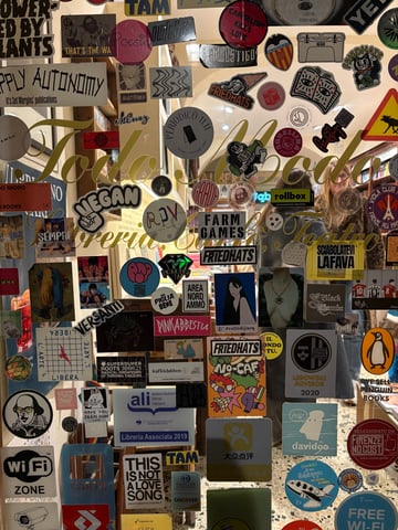 A dense and vibrant collage of various stickers and decals layered onto a transparent glass shop window. The collection forms a complex visual tapestry of logos, illustrations, and slogans that partially obscures the interior of a bookstore.. Subject: A cartoon coffee character mascot for 'Friedhats'. Style: Illustrated cartoon. Details: Bold black outlines with a playful, expressive coffee cup character, appearing multiple times in different variations across the glass.. Shape: Circular. Setting: Indoor bookstore/cafe environment. Surface: A glass shopfront window or entrance door for a literary cafe.. Colors: #000000 (Black), #FFFFFF (White), #FFD700 (Gold), #C51F2D (Red), #1E40AF (Deep Blue). Composition: A flat, frontal perspective focusing on the two-dimensional plane of the sticker collage.. The glass serves as a communal canvas where commercial branding, political messages, and independent art coexist. This layering represents a 'visual noise' that characterizes urban hubs, reflecting the diverse cultural influences and the identity of the establishment as a social crossroads.