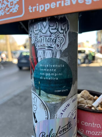 A close-up shot of a grey metal pole densely covered with various street stickers in an urban environment. The central focus is a circular teal sticker featuring evocative Italian text superimposed over a dark, moody landscape.