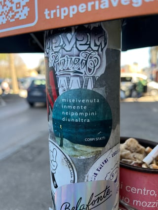A close-up shot of a grey metal pole densely covered with various street stickers in an urban environment. The central focus is a circular teal sticker featuring evocative Italian text superimposed over a dark, moody landscape.
